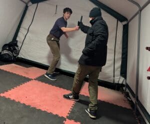 **Alt text:**

> Two men demonstrating a self-defence technique on interlocking gym mats inside a training marquee. One man intercepts and controls the weapon arm of an attacker holding a training blade, illustrating the "defanging the snake" concept.