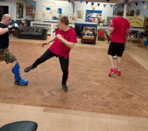 A practitioner in a red t-shirt executes a low interception kick toward a partner wearing a shin guard and boxing gloves, demonstrating a Jeet Kune Do technique.